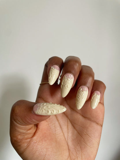 A hand showcasing medium almond-shaped beige false nails with glossy finish and ornate 3D designs.
